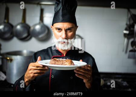 Chef professionista con una barba lunga indossando l'uniforme nera, che tiene un piatto con una deliziosa bistecca alla griglia nella cucina del ristorante Foto Stock