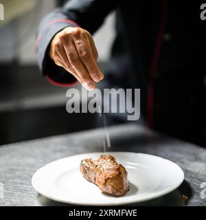 Lo chef aggiunge un tocco finale a un succulento piatto di bistecca, spolverando sale su un piatto bianco. Primo piano cattura le mani esperte in azione, mostrando arte culinaria Foto Stock