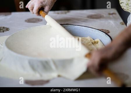 La nonna vecchia mette l'impasto arrotolato su una teglia con un mattarello. Donna anziana che prepara la tradizionale torta balcanica Foto Stock