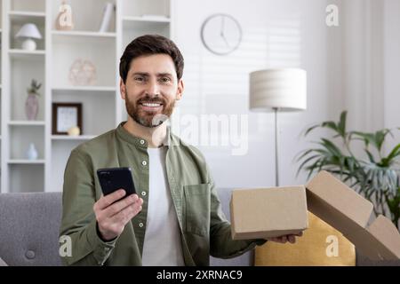 Uomo sorridente che tiene il telefono e apre una scatola di cartone nel soggiorno. Concetto di shopping online, consegna, casa e tecnologia. Emozioni positive e uno stile di vita moderno catturati al chiuso. Foto Stock