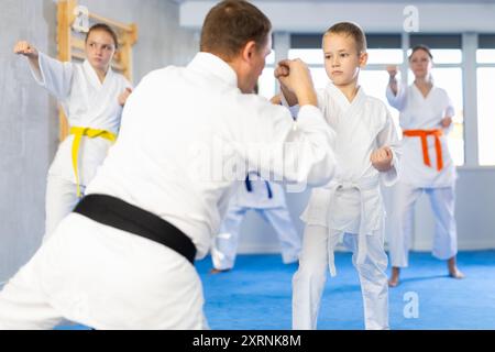 Ragazzo con la sua famiglia in kimono e cinture colorate che pratica karate con pugni durante la lezione di arti marziali di gruppo in palestra, accompagnato da un allenatore Foto Stock