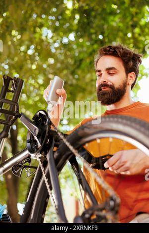 Uomo sano e energico che applica grasso sulla catena della bicicletta e ruota di regolazione all'esterno. Sportivo maschile caucasico che controlla e mantiene la bicicletta per il ciclismo estivo annuale. Foto Stock