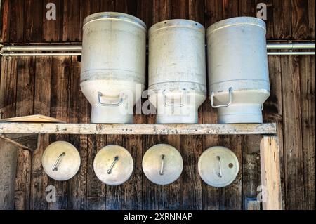 Contenitori del latte in metallo impilati e coperchi appesi su una parete di legno di un cottage alpino. Foto Stock