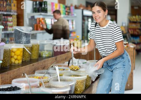 La ragazza sceglie le olive marinate nel supermercato, raccogliendole dal secchio con mestolo in un sacchetto di plastica Foto Stock