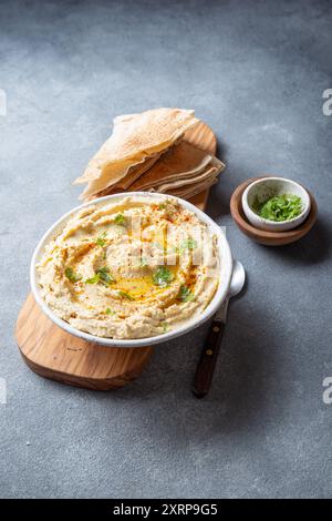 Ciotola di hummus cremoso guarnita con olio d'oliva ed erbe aromatiche viene servita con pane pita su un asse di legno. Accanto al è presente un piccolo recipiente di modanatura Foto Stock