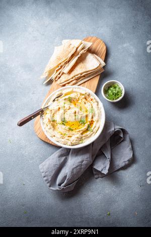 Ciotola di hummus cremoso guarnita con olio d'oliva ed erbe aromatiche viene servita con pane pita su un asse di legno. Accanto al è presente un piccolo recipiente di modanatura Foto Stock