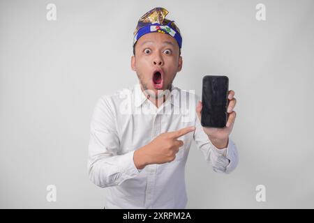 Sorpreso giovane balinese che indossa una camicia bianca e un copricapo tradizionale con un'espressione scioccata sul viso mentre punta al suo smartphone, presentin Foto Stock