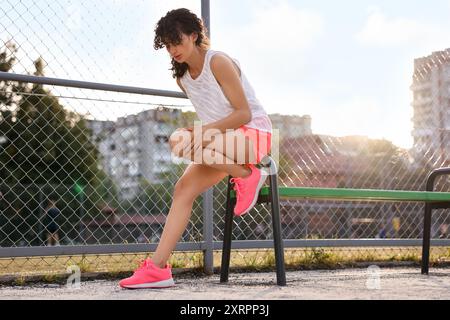 Donna sportiva con lesioni alle gambe che soffre di dolore al ginocchio vicino alla panchina dello stadio Foto Stock