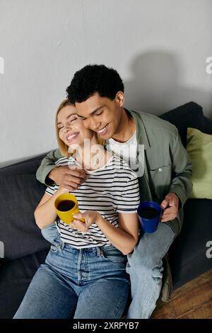 Una giovane coppia interrazziale si rilassa su un divano in un appartamento moderno, condividendo un caffè e un momento accogliente insieme. Foto Stock