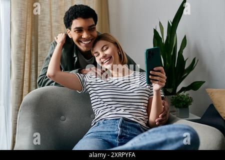 Una giovane coppia interrazziale ride insieme in un appartamento moderno tenendo in mano uno smartphone. Foto Stock