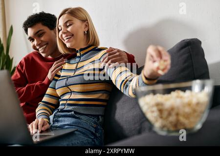 Una coppia felice e multiculturale si diverte a trascorrere una notte in compagnia, condividendo una risata mentre si guarda un film su un notebook. Foto Stock