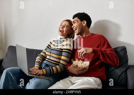 Una felice coppia interrazziale ride insieme mentre guardano un film su un computer portatile nel loro accogliente appartamento moderno. Foto Stock