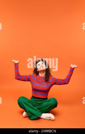 Donna dai capelli ricci in posa con le braccia alzate seduta su uno sfondo arancione brillante, vestita con maglione rosso e viola con pantaloni verdi. Foto Stock