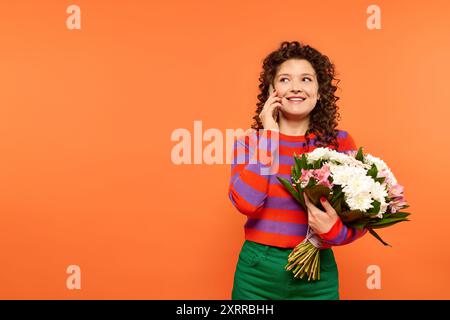 Una giovane donna con i capelli ricci sorride brillantemente mentre tiene un mazzo di fiori e parla al telefono su uno sfondo arancione. Foto Stock