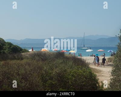 Panorama orizzontale di un sentiero attraverso lussureggianti cespugli che conduce alla spiaggia di Nodu Pianu in Sardegna con ombrelloni, yacht e colline in lontananza Foto Stock