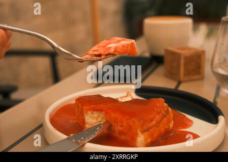 Una persona sta mangiando un pezzo di torta con una forchetta. La torta è ricoperta da una salsa di fragole rosse Foto Stock