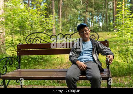 Un ragazzino che indossa occhiali e un cappello blu siede su una panchina del parco in una foresta Foto Stock