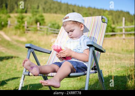 Il bambino siede comodamente su una sedia pieghevole in un ambiente all'aperto erboso, giocando intensamente con un giocattolo rosa. Il bambino indossa un cappellino all'indietro, una camicia a motivi geometrici e pantaloncini corti. Splendido paesaggio verde sullo sfondo. Foto Stock