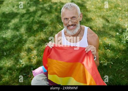 Un gay maturo con una barba grigia e tatuaggi sorride mentre tiene una bandiera arcobaleno all'aperto. Foto Stock