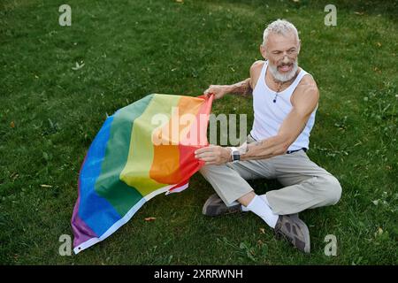 Un gay maturo con tatuaggi e barba grigia tiene una bandiera arcobaleno mentre si siede sull'erba. Foto Stock