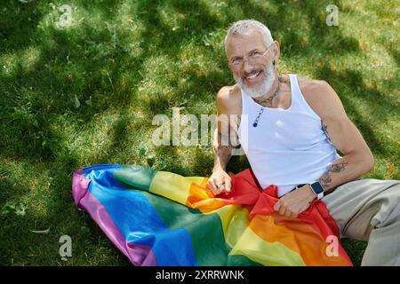 Un gay maturo con tatuaggi e barba grigia sorride e tiene in mano una bandiera arcobaleno all'aperto nell'erba. Foto Stock