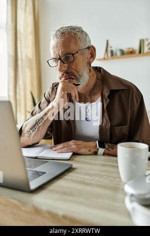 Un uomo gay maturo con capelli grigi e tatuaggi lavora in remoto da casa, concentrandosi sul suo notebook e notebook. Foto Stock