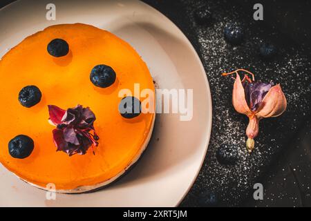 Dessert o torta con strati di cioccolato, crema di cagliata, gelatina di mango, decorata con cibo fiori secchi su un piatto bianco su sfondo nero. Originale Foto Stock