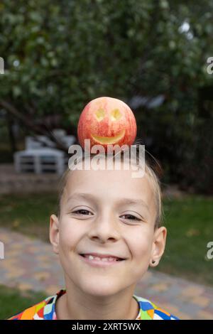 faccia di un ragazzo carino di 7-8 anni, una mela allegra con un sorriso intagliato giace sulla testa. Divertente pubblicità per la frutta fresca. Concetto di alimentazione sana. Foto Stock