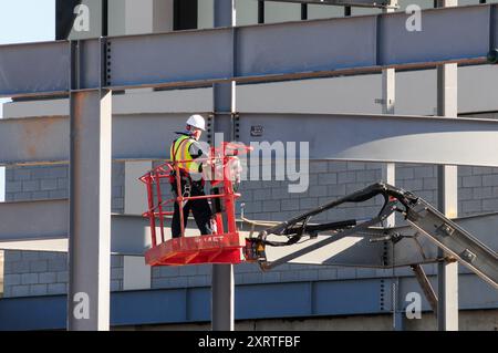 Il lavoratore edile con elmetto e giubbotto di sicurezza si trova sulla piattaforma di un veicolo di sollevamento aereo in un edificio in acciaio e mattoni in costruzione Foto Stock