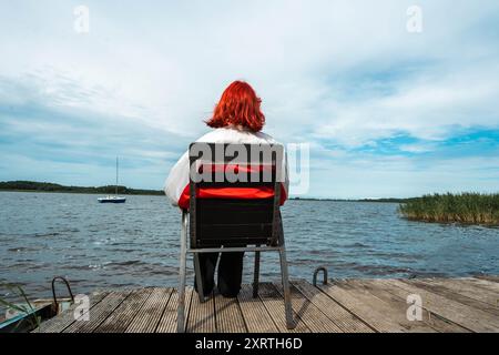 Una ragazza irriconoscibile con i capelli rossi è seduta su una sedia vicino al lago e guarda l'acqua. Generazione Z Foto Stock