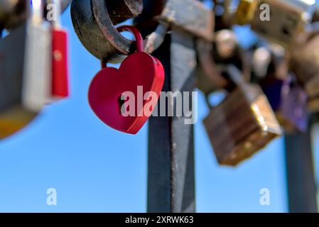 Lucchetto rosso a forma di cuore che simboleggia l'amore, attaccato ad una ringhiera tra gli altri lucchetti in una giornata di sole. Foto Stock