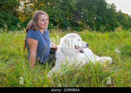 Bella giovane donna e il suo carino grande cane bianco dei grandi Pirenei che giace in un prato erboso alto, godendosi del tempo insieme. Proprietario di animali domestici, amore per animali domestici, patatine fritte per cani Foto Stock