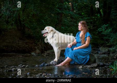 La bella giovane donna e il suo grande cane bianco dei grandi Pirenei si divertono insieme, rinfrescandosi in un ruscello durante le calde giornate estive. Proprietario di animali domestici, lov Foto Stock