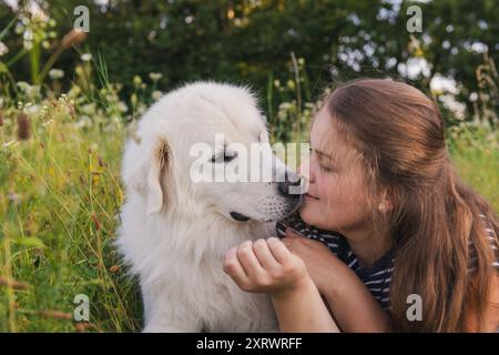 Bella giovane donna che baciava il suo carino grande cane bianco dei grandi Pirenei steso su un prato erboso alto, godendosi del tempo insieme. Proprietario di animali domestici, amore per animali, fr. Cane Foto Stock