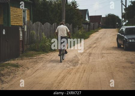 Il nonno pedala su una strada sterrata nel villaggio. Un vecchio in bicicletta Foto Stock