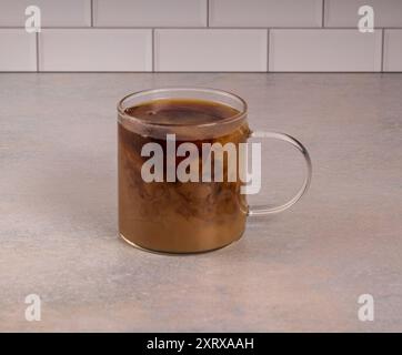 La cremosa gira in una tazza calda di caffè in una tazza di bicchiere seduta su un bancone di colore rosa con sfondo bianco con piastrelle metropolitane. La condensa è forma Foto Stock