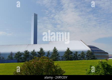 Alto camino bianco sopra un edificio industriale con tetto curvo, adagiato contro il cielo blu e le nuvole bianche. Fabbrica di knoll erboso con alberi davanti Foto Stock