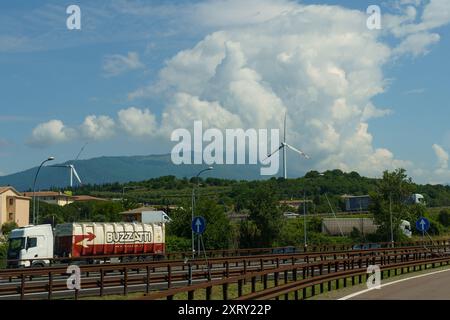 Ossenigo, Italia - 8 giugno 2023: Le turbine eoliche sovrastano le colline ondulate, mescolandosi con le spettacolari formazioni nuvolose mentre i veicoli attraversano la panoramica roa Foto Stock