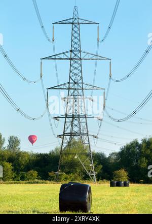 Piloni e pallone di passaggio a Kennington Meadows. Sono sempre stato affascinato dai nostri piloni britannici - quelle che in gran parte del mondo sarebbero chiamate torri di trasmissione. Marciando attraverso il paesaggio, hanno un impatto visivo così forte e sorprendente. Si trova a Kennington Meadows, tra Oxford e Abingdon, e una mongolfiera si aggira maestosamente nelle prime ore del mattino. Ma quali sono esattamente le misteriose luccicanti borse nere? Sfortunatamente, non sono escrementi alieni giganti. In realtà contengono insilato di balle per l'alimentazione del bestiame; per prima cosa, prendete il tradizionale rotolo di fieno e avvolgetelo in plastica A. Foto Stock