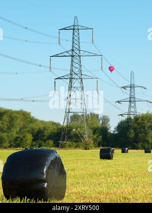 Piloni e pallone di passaggio a Kennington Meadows. Sono sempre stato affascinato dai nostri piloni britannici - quelle che in gran parte del mondo sarebbero chiamate torri di trasmissione. Marciando attraverso il paesaggio, hanno un impatto visivo così forte e sorprendente. Si trova a Kennington Meadows, tra Oxford e Abingdon, e una mongolfiera si aggira maestosamente nelle prime ore del mattino. Ma quali sono esattamente le misteriose luccicanti borse nere? Sfortunatamente, non sono escrementi alieni giganti. In realtà contengono insilato di balle per l'alimentazione del bestiame; per prima cosa, prendete il tradizionale rotolo di fieno e avvolgetelo in plastica A. Foto Stock