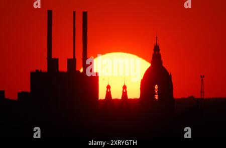 Hannover, Germania. 12 agosto 2024. Il sole tramonta dietro l'impianto di cogenerazione di Linden e il nuovo Municipio. La foto è stata scattata da otto chilometri di distanza dal municipio di Kronsberg con un teleobiettivo estremo. Credito: Julian Stratenschulte/dpa/Alamy Live News Foto Stock