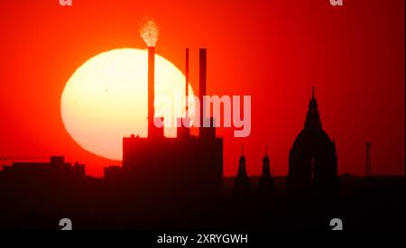 Hannover, Germania. 12 agosto 2024. Il sole tramonta dietro l'impianto di cogenerazione di Linden e il nuovo Municipio. La foto è stata scattata da otto chilometri di distanza dal municipio di Kronsberg con un teleobiettivo estremo. Credito: Julian Stratenschulte/dpa/Alamy Live News Foto Stock