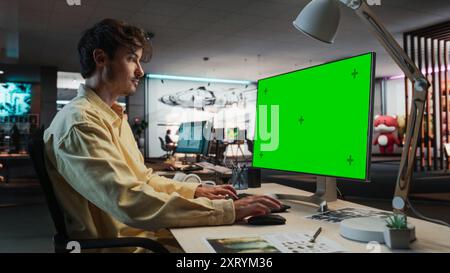 Uomo caucasico che utilizza un computer desktop con Mock-up Green Screen Chromakey. Artista concettuale maschile che lavora in Game Design Startup Variety Office, creando un'azione di gioco coinvolgente per New Survival Video Game. Foto Stock