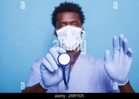 Ritratto dell'infermiera nera che utilizza lo stetoscopio per ascoltare i battiti cardiaci durante il controllo cardiologico. Uomo con strumento medico, indossa maschera e guanti per proteggere da epidemia di coronavirus. Foto Stock