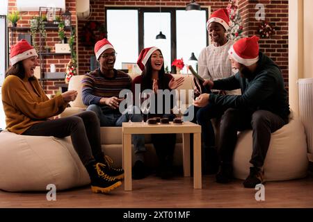 Allegri colleghi di babbo natale che versano spumante mentre festeggiano le vacanze invernali in un luogo di lavoro decorato. Dipendenti diversi felici che si riuniscono intorno al tavolo con bevande alcoliche alla vigilia di Natale Foto Stock
