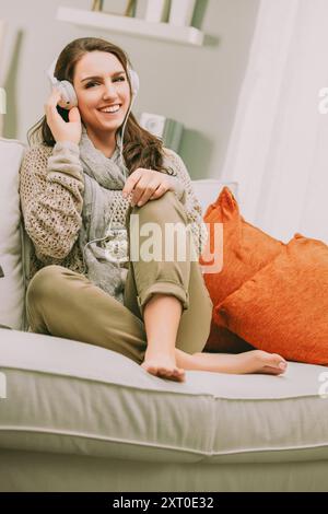 donna con lunghi capelli marroni seduto comodamente su un divano, sorridendo mentre ascolta la musica attraverso le cuffie di grandi dimensioni. Indossato con un comodo maglione e sciarpa, Foto Stock