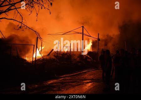 Atene, Grecia, 12 agosto 2024. Un magazzino di legname è in fiamme mentre la gente si riunisce fuori nel sobborgo di Gerakas, capitale greca, durante un incendio che ha bruciato case. Un incendio che scoppiò a Varnavas l'11 agosto continuò a infuriare nell'Attica orientale il 12 agosto, si diffuse e si diffuse su un fronte che si estendeva per più di 20 chilometri. Diversi paesi hanno risposto positivamente alla richiesta delle autorità greche di assistenza da parte del meccanismo di protezione civile dell'UE (EUCPM) per combattere l'infuria degli incendi. Crediti: Dimitris Aspiotis/Alamy Live News Foto Stock