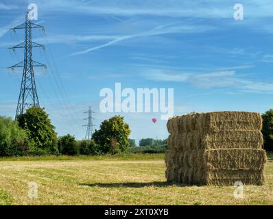 E' subito dopo l'ora del raccolto. Visto in un campo di grano appena fuori dal mio villaggio di casa, Radley, Oxfordshire, questo gigantesco pagliaio sembra aver trascendito il suo nome; forse questo imponente edificio potrebbe essere chiamato "grattacielo di fieno" o "grattacielo di fieno"...? Contrasta piacevolmente con gli onnipresenti piloni elettrici che coprono questa parte rurale dell'Oxfordshire. Nel frattempo, una mongolfiera passa davanti in questa bella e soleggiata mattinata. Foto Stock