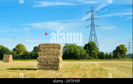 E' subito dopo l'ora del raccolto. Visto in un campo di grano appena fuori dal mio villaggio di casa, Radley, Oxfordshire, questo gigantesco pagliaio sembra aver trascendito il suo nome; forse questo imponente edificio potrebbe essere chiamato "grattacielo di fieno" o "grattacielo di fieno"...? Contrasta piacevolmente con gli onnipresenti piloni elettrici che coprono questa parte rurale dell'Oxfordshire. Nel frattempo, una mongolfiera passa davanti in questa bella e soleggiata mattinata. Foto Stock