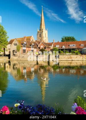 Una bella vista del Tamigi ad Abingdon, nelle prime ore di una mattina d'estate. Siamo sulla riva sud del fiume, guardando dall'altra parte verso la chiesa anglosassone di Sant'Elena da cui prende il nome il molo, passando davanti a una casa galleggiante ormeggiata e ricoperta di fiori in primo piano. Foto Stock